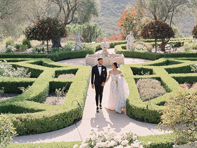 A newlywed couple holding hands and walking through an elegant garden maze at a Southern California wedding venue. The groom is dressed in a classic black tuxedo, while the bride wears an off-the-shoulder lace gown with a long train. Statues and manicured hedges line the path under a sunny sky.