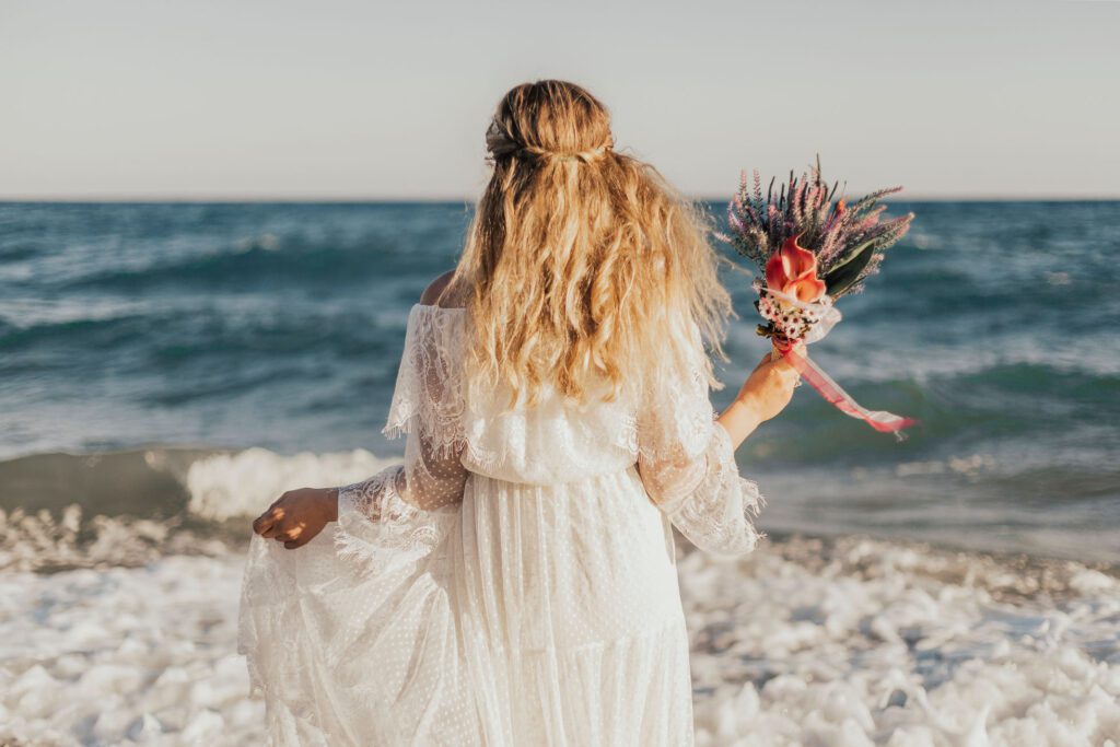 A woman clad in a lace beach wedding dress stands with her back to the camera, holding a bouquet of pink and purple flowers, with the ocean waves gently crashing onto the shore in the background at a San Diego beach wedding. 