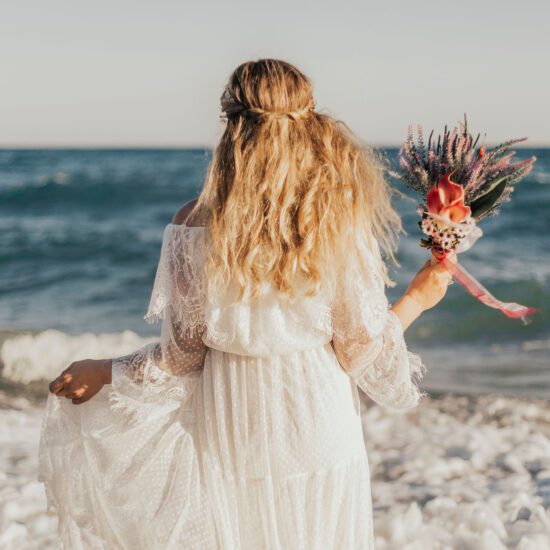 A woman clad in a lace beach wedding dress stands with her back to the camera, holding a bouquet of pink and purple flowers, with the ocean waves gently crashing onto the shore in the background.