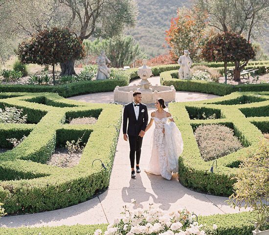 A newlywed couple holding hands and walking through an elegant garden maze at a Southern California wedding venue. The groom is dressed in a classic black tuxedo, while the bride wears an off-the-shoulder lace gown with a long train. Statues and manicured hedges line the path under a sunny sky.